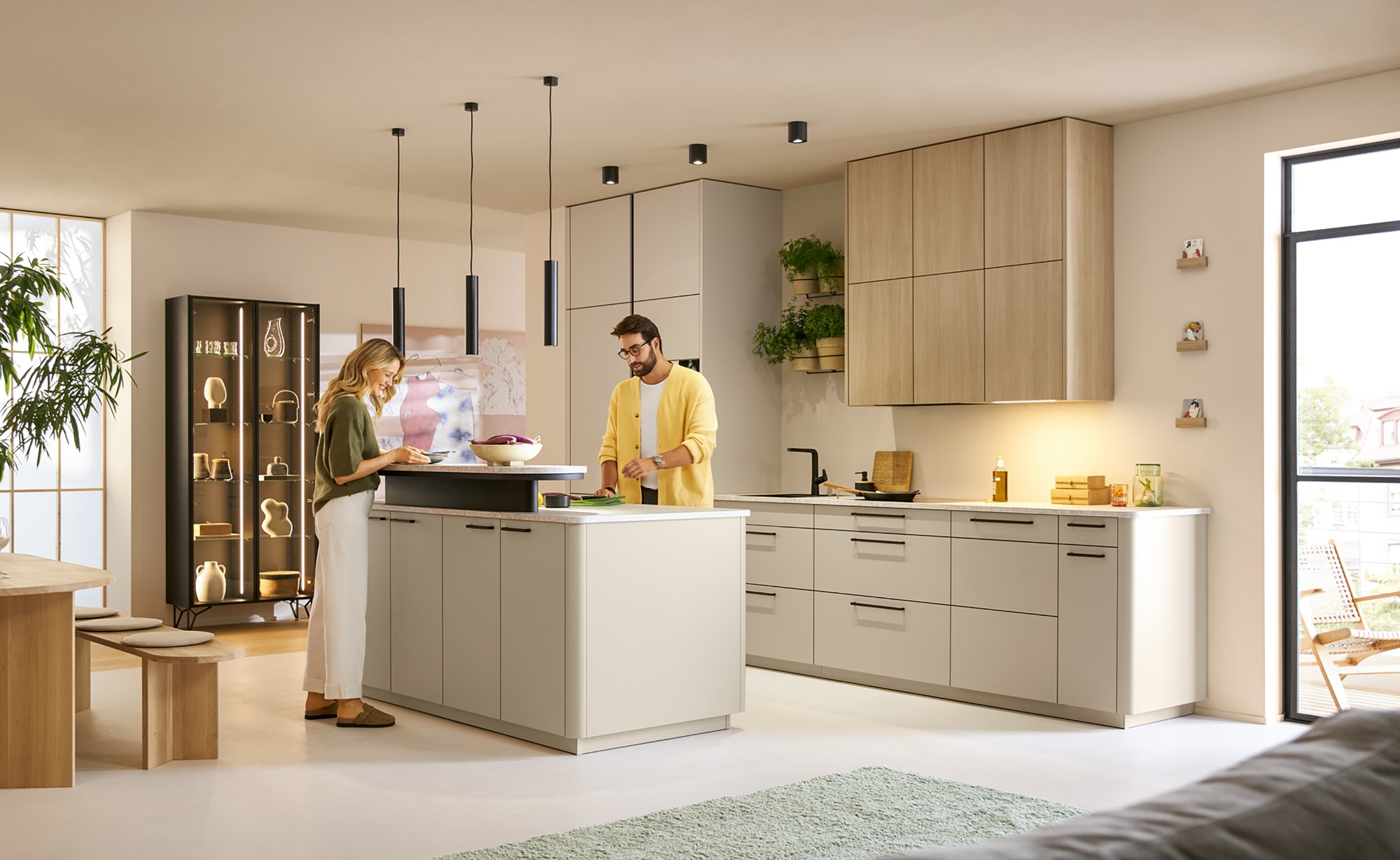 Moderne Küche mit zwei Personen, die gemeinsam am Kochfeld arbeiten. Helle Farben und Pflanzen.