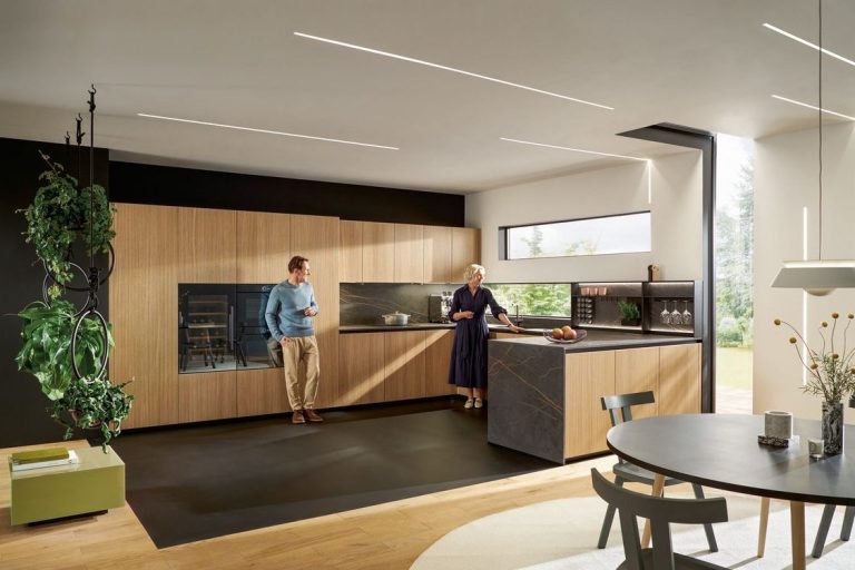 Küchen Moderne Küche mit Holzschränken, Tisch und zwei Personen beim Arbeiten. Fronten sind in Holz, die Arbeitsplatte in Marmornachbildung dunkel.